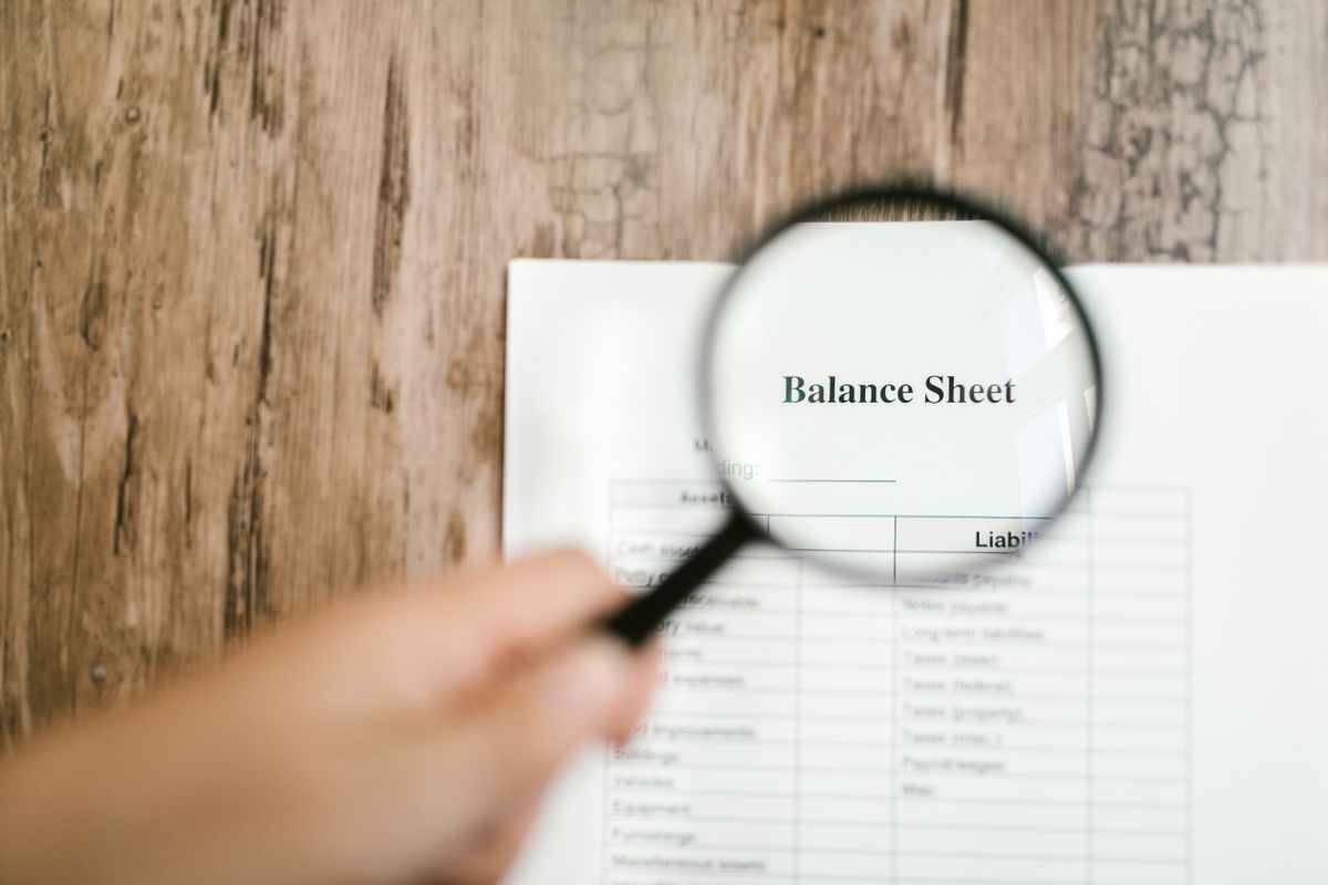 A hand holding a magnifying glass over financial documents, representing the codebase audit needed to find every dynamic API call before it breaks a static export build
